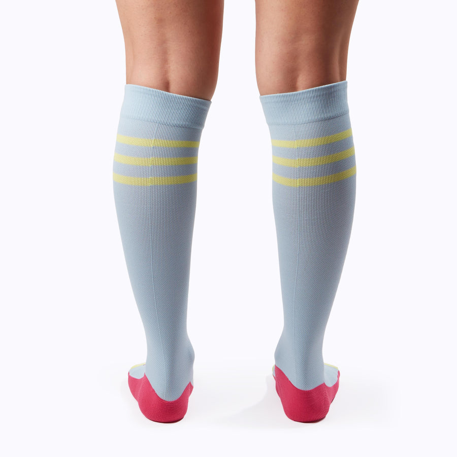 Back  view of a pair of legs wearing varstity stripes knee high compression socks in sky blue and pear