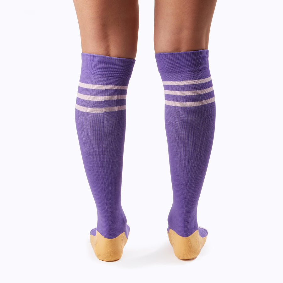 Back  view of a pair of legs wearing varstity stripes knee high compression socks in violet and bubblegum